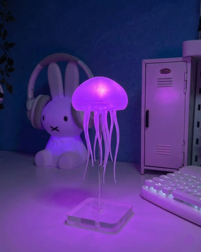 Lámpara de medusas LumaJelly™ en color púrpura iluminando un escritorio con estética gamer, teclado mecánico blanco y accesorios modernos.